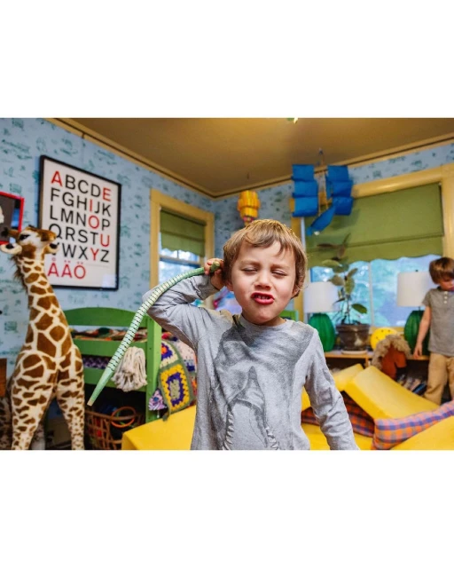 A young boy makes a face indoors while holding a green hose up to his ear, with another child and a toy giraffe visible in the background.