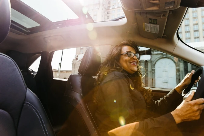 Smiling woman with glasses driving a car, looking through the windshield, with a city building visible in the background.