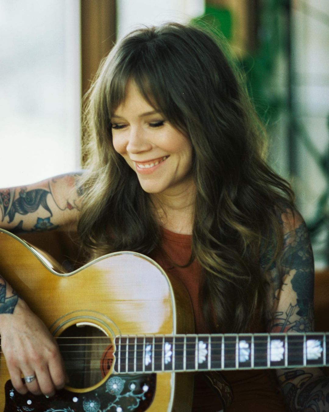Smiling woman with tattoos plays acoustic guitar indoors. Portrait captures natural light, musical performance, and detailed instrument.