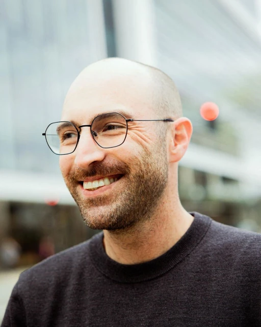 Close-up of a smiling, bald adult man wearing glasses and a dark sweater, set against a blurred architectural background.