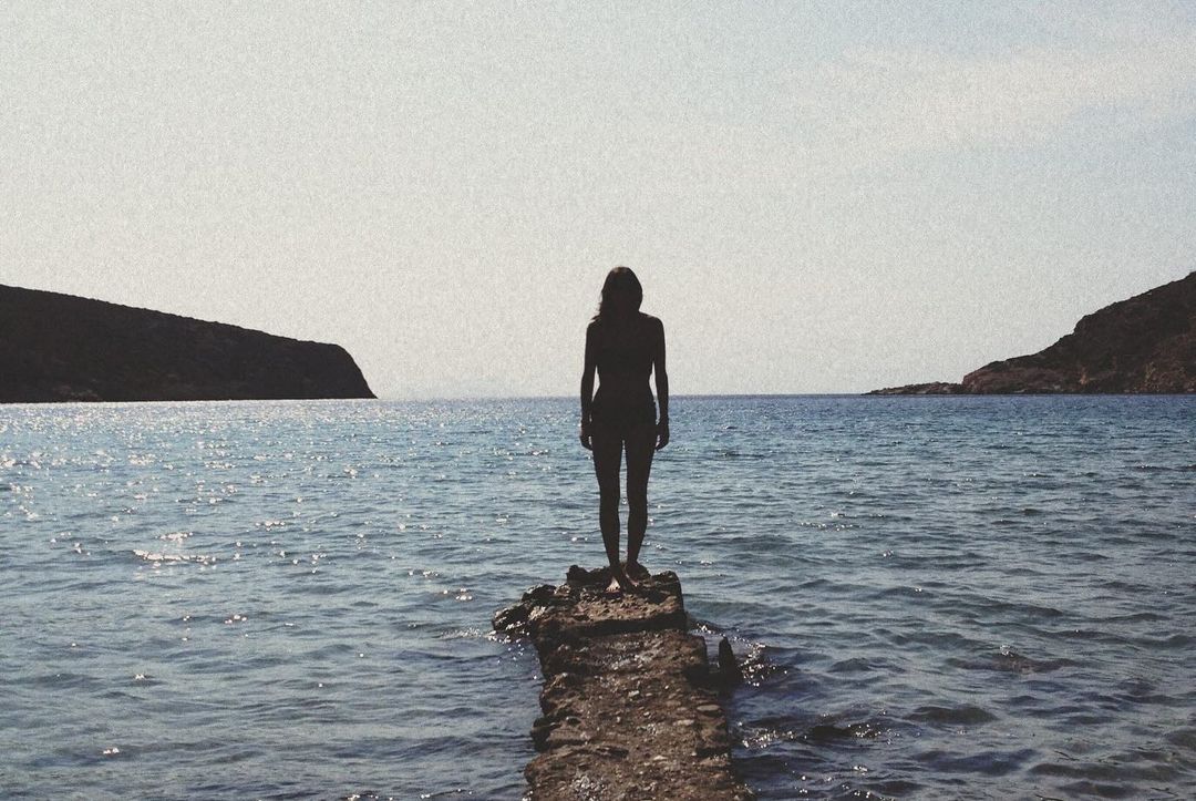 Silhouette of a woman standing on a rocky outcrop in the sea, with hills in the distance under a bright sky, creating a serene seascape.