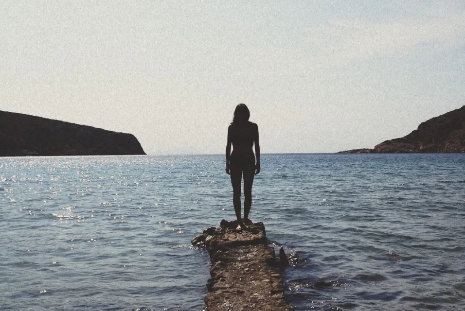 Silhouette of a woman standing on a rocky outcrop in the sea, with hills in the distance under a bright sky, creating a serene seascape.