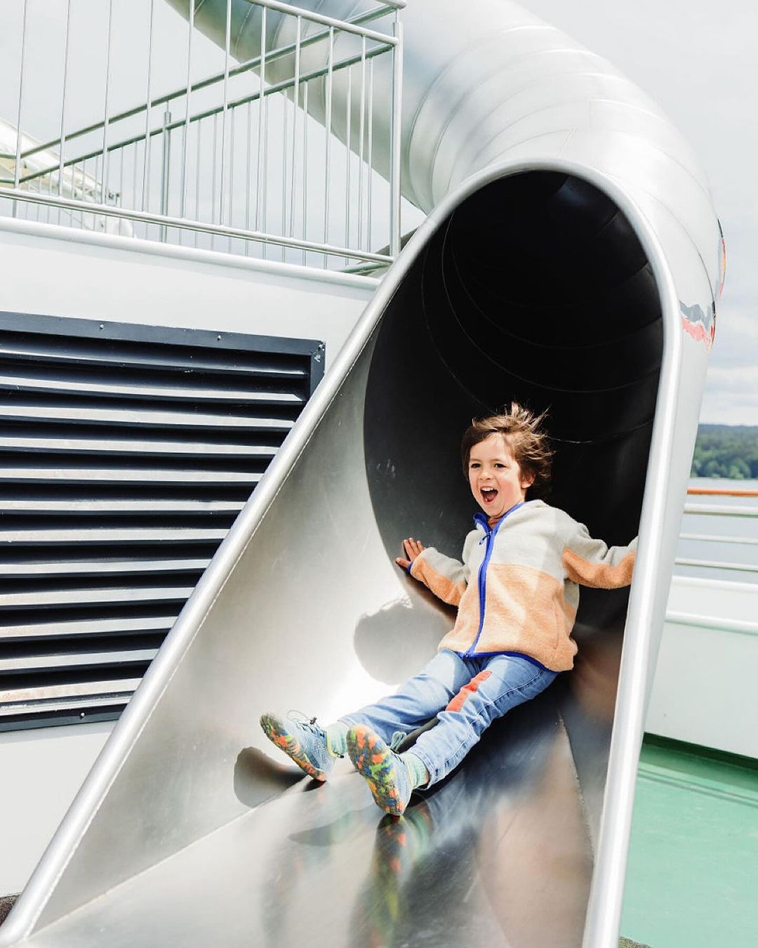 Excited child with open mouth in blue jeans and jacket sliding down an outdoor metal slide with green ground in bright sunlight.