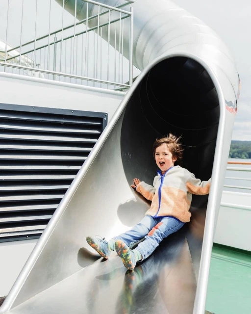 Excited child with open mouth in blue jeans and jacket sliding down an outdoor metal slide with green ground in bright sunlight.