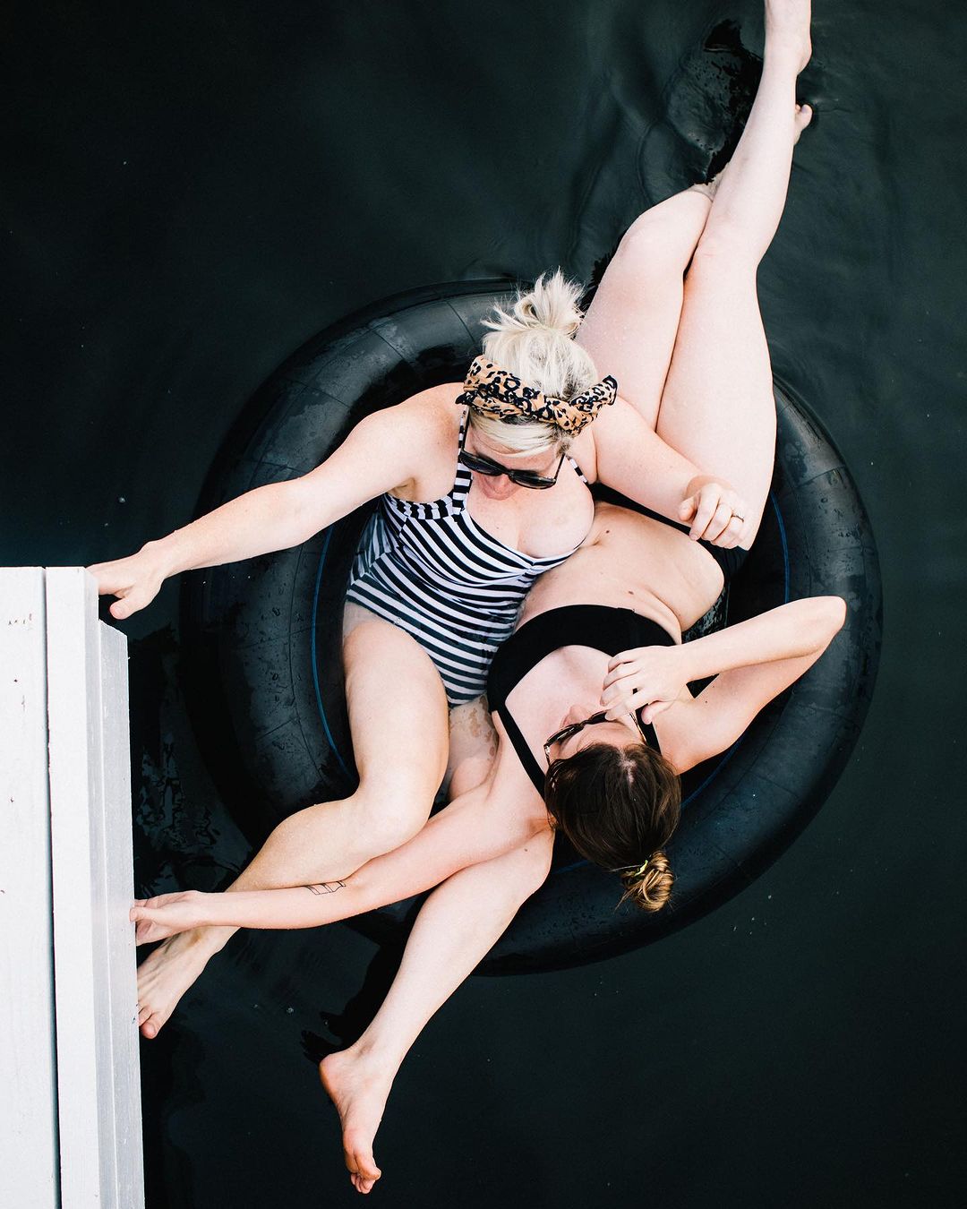 Two women in swimsuits relax in a black inner tube on dark water next to a white dock, captured from an overhead angle in sunny daylight.