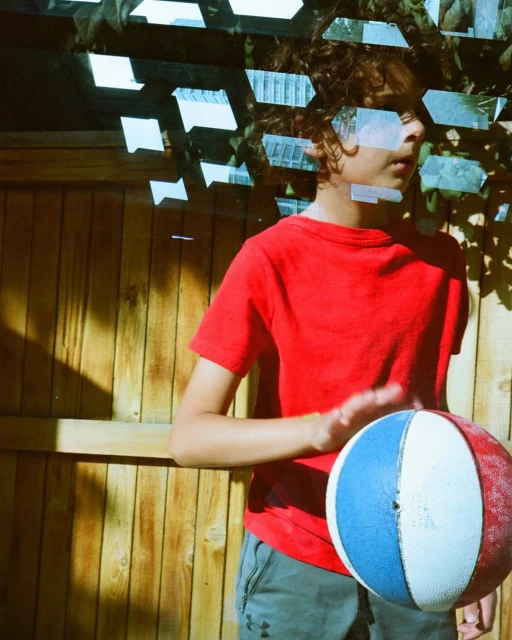 A kid plays with a red, white, and blue basketball in a backyard with a wooden fence, dappled light, and artistic light effects.