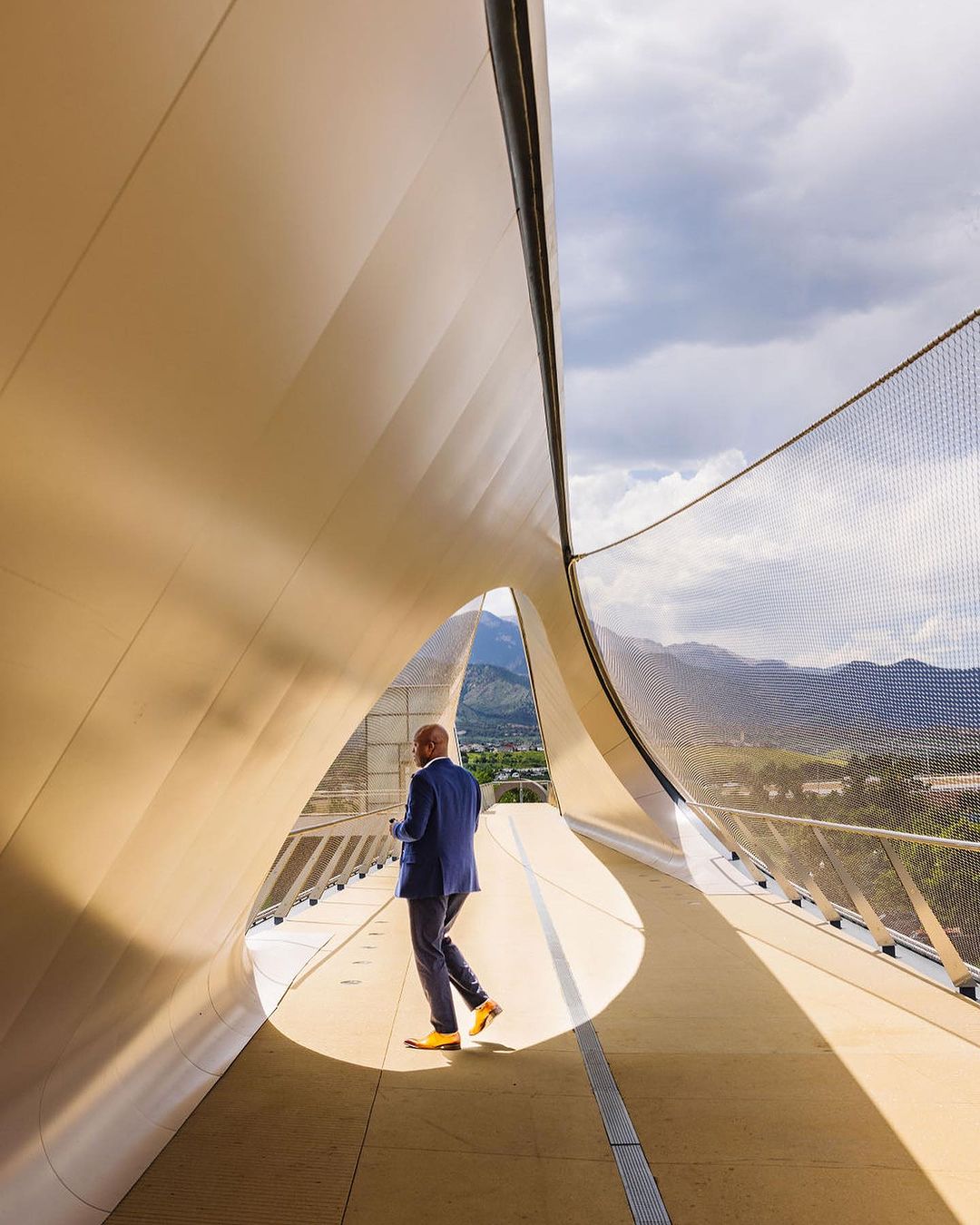 Adult male in blue suit walks on modern pedestrian bridge with yellow shoes; background view of mountains and sky with clouds.