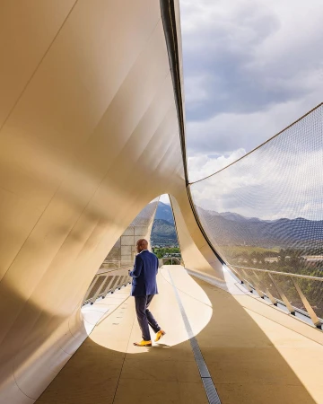 Adult male in blue suit walks on modern pedestrian bridge with yellow shoes; background view of mountains and sky with clouds.