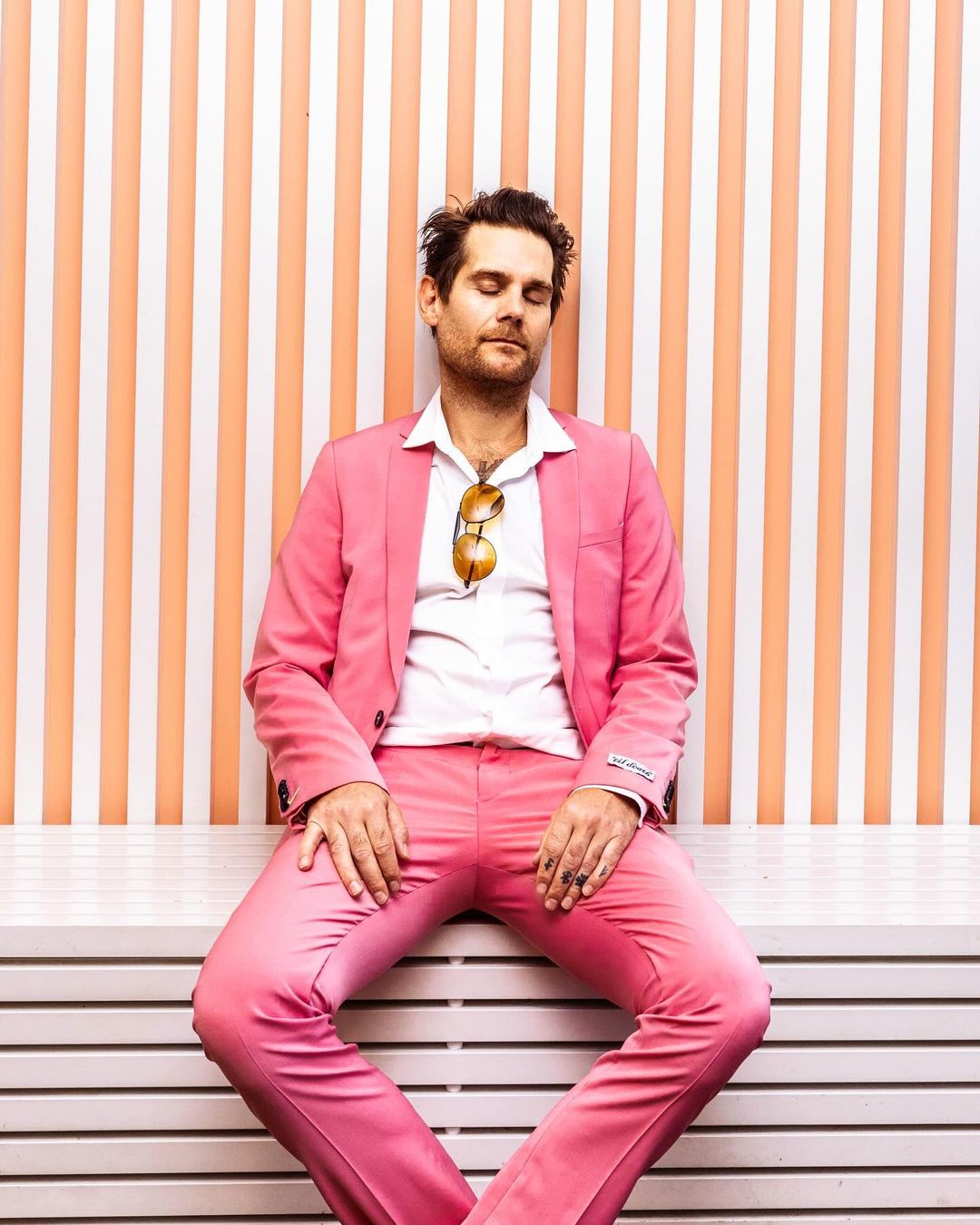 Man in a pink suit with sunglasses posing against a peach and white striped wall with his eyes closed, sitting down on horizontal slats.