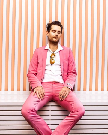 Man in a pink suit with sunglasses posing against a peach and white striped wall with his eyes closed, sitting down on horizontal slats.