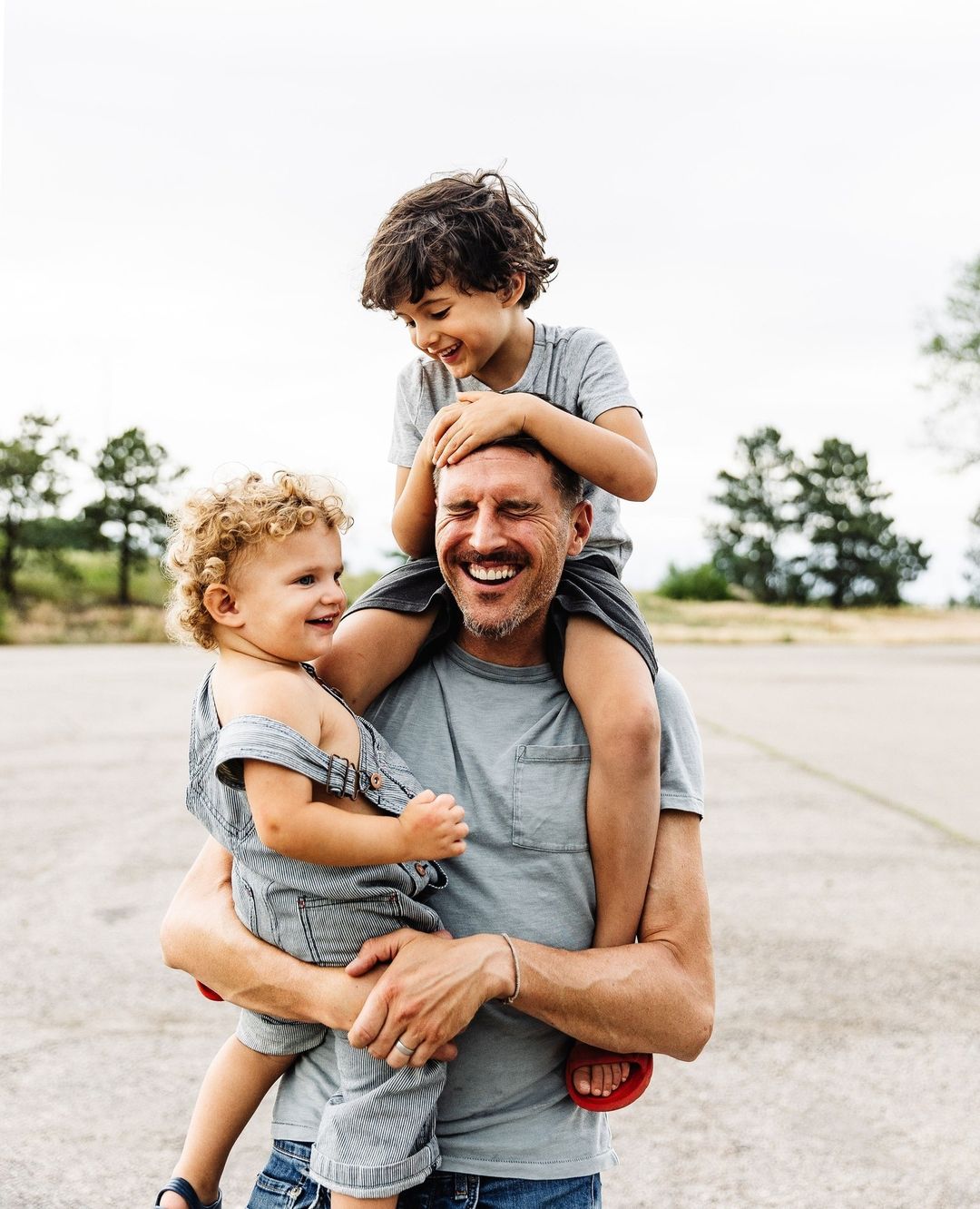 Father carries two young children on shoulders outside. Kids are smiling. One child wears a grey outfit, the other wears blue overalls.