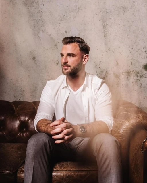 Man sits on a leather couch in front of a textured grey wall. He is wearing a white shirt and grey pants. He looks off to the side.