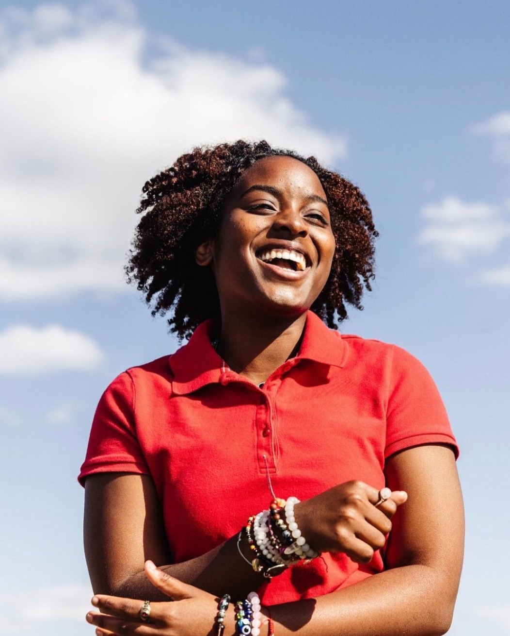 Portrait of a smiling young Black woman outdoors wearing a red polo shirt, multiple bracelets, and rings, set against a blue sky and clouds.