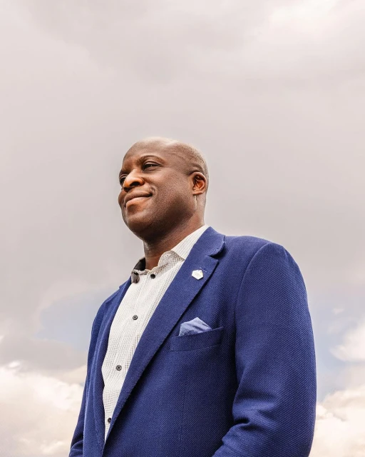 Portrait of a dark-skinned adult male in a blue blazer with a white dress shirt and a light-colored cloudy sky background.