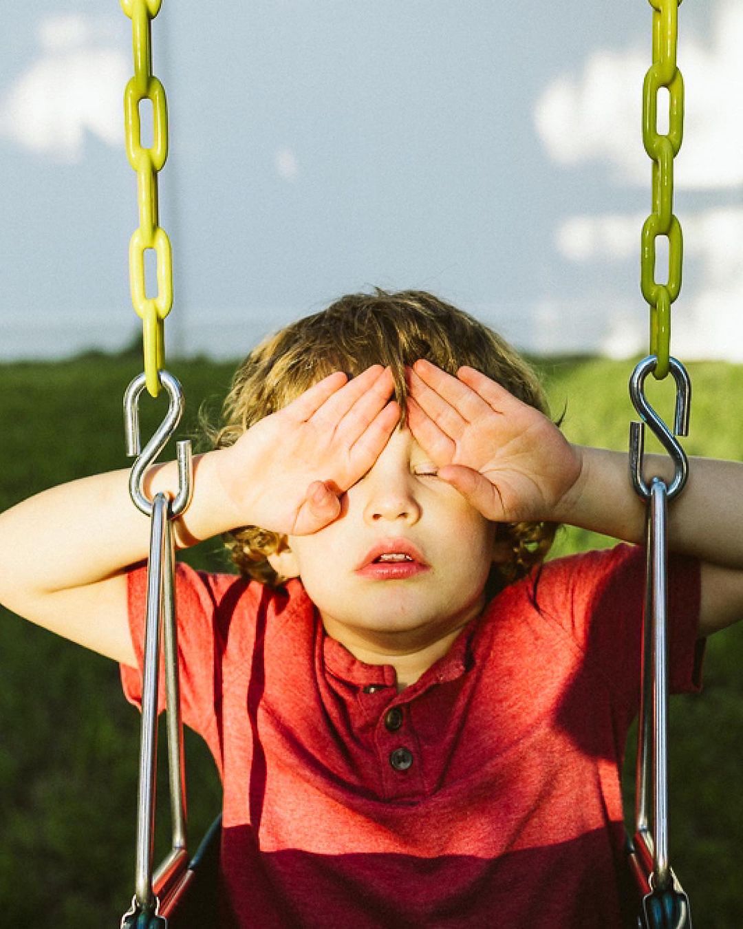 Boy with curly brown hair in a red shirt sits on a yellow swing with hands covering eyes outside on a sunny day with a green lawn.