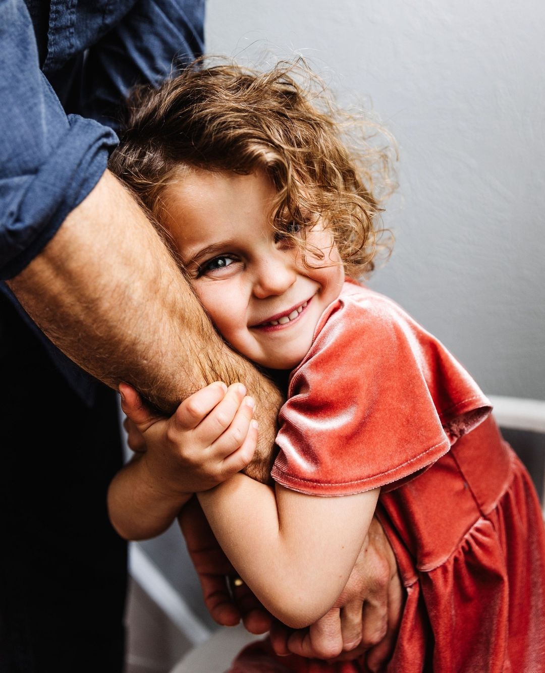 A smiling young girl in a pink dress hugs an adult's arm. The child has curly hair and blue eyes, and she is indoors with natural light.