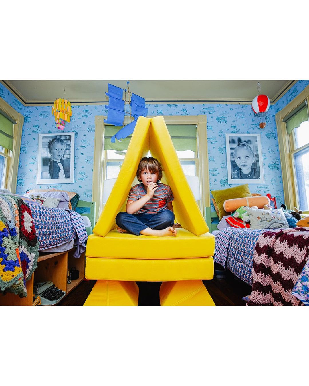 Boy sits inside of a yellow foam play structure, in a bedroom setting with blue patterned wallpaper, and two beds visible.