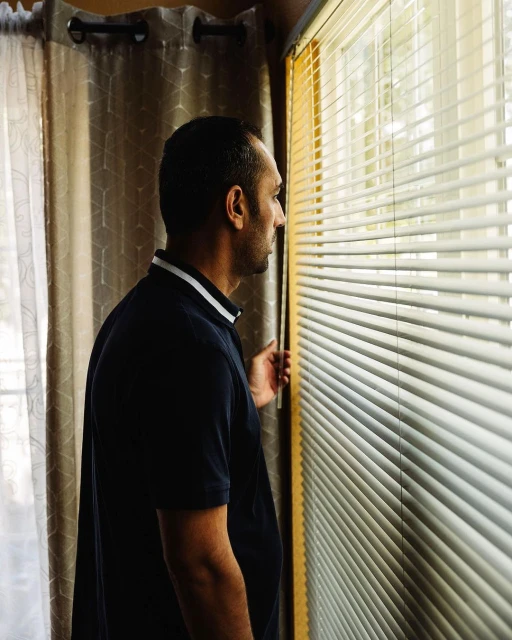 Adult male in a dark polo shirt looking out the window through closed blinds in an indoor setting with curtains in the background.