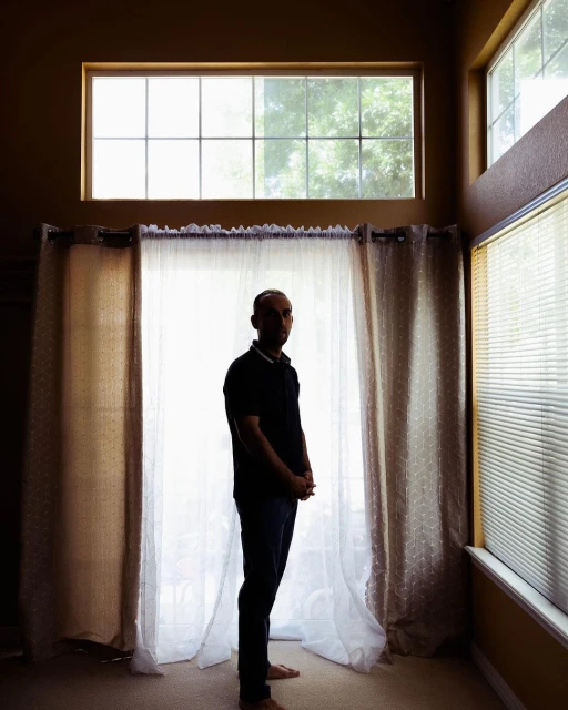 Full shot of an adult male in a dark shirt and jeans stands in front of a large window with sheer curtains in a high-ceilinged room.