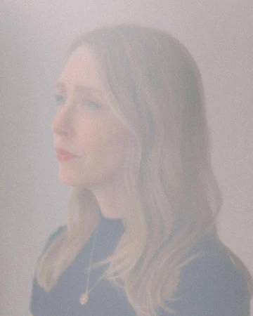Portrait of a woman with long wavy blonde hair wearing a dark top, captured through a hazy or frosted glass filter.