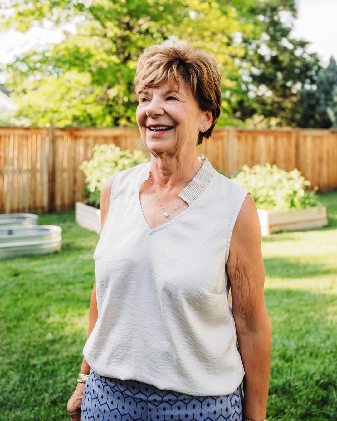 Outdoor portrait of a smiling mature woman with short hair wearing a light-colored top and patterned pants, standing in a backyard.