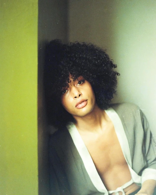 Portrait of a young adult with dark curly hair, wearing a robe, posing against a green wall in a naturally lit indoor setting.