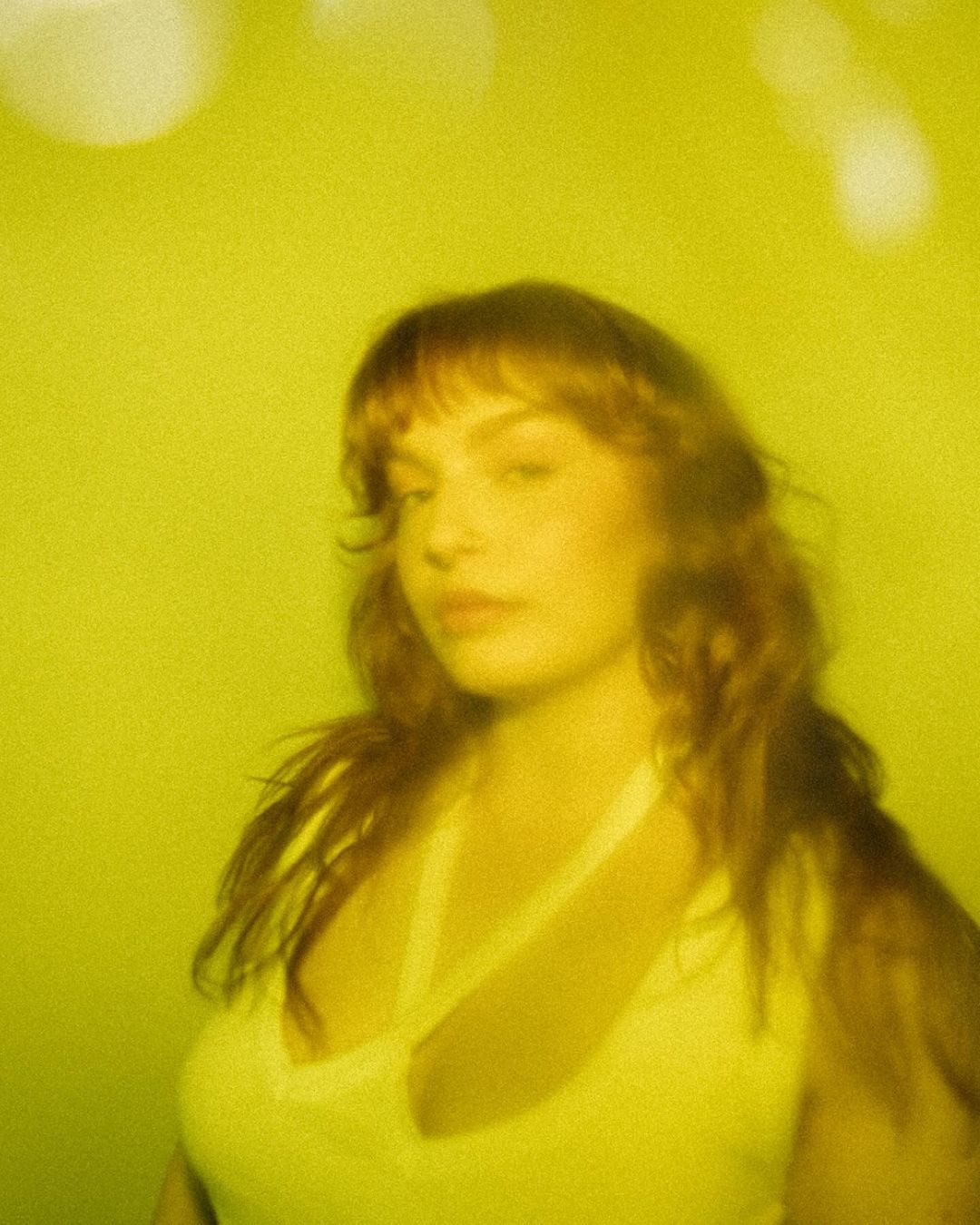 Portrait of an adult female with auburn hair and bangs, wearing a white top, posing in a blurred, golden-yellow monochrome environment.