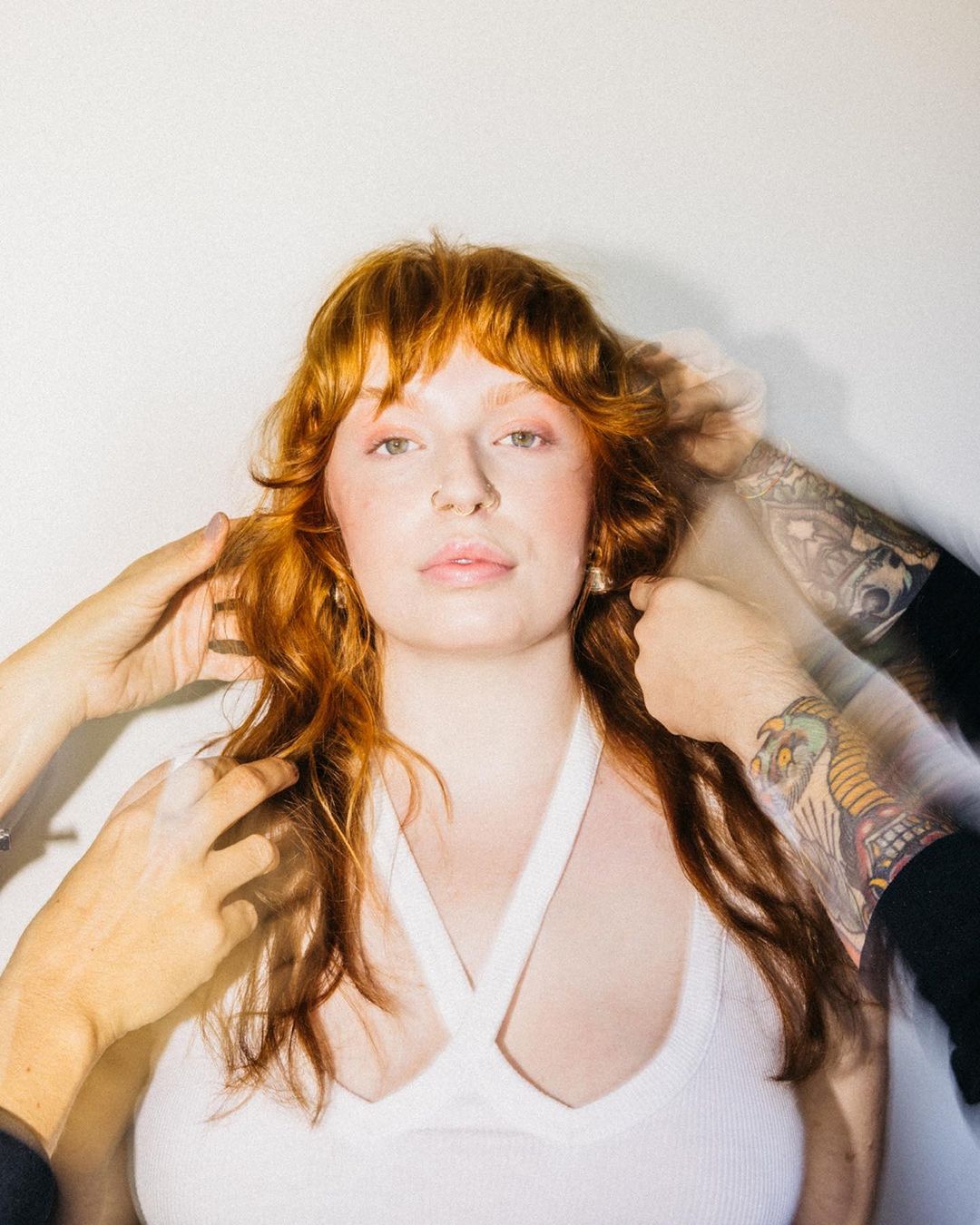 Portrait of a red-haired woman with hands framing her face in a studio setting, wearing a white tank top and gold earrings.