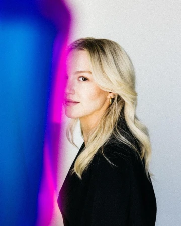 Blonde woman wearing a black top with vibrant blue and pink lighting against a white background by Stephanie Bassos