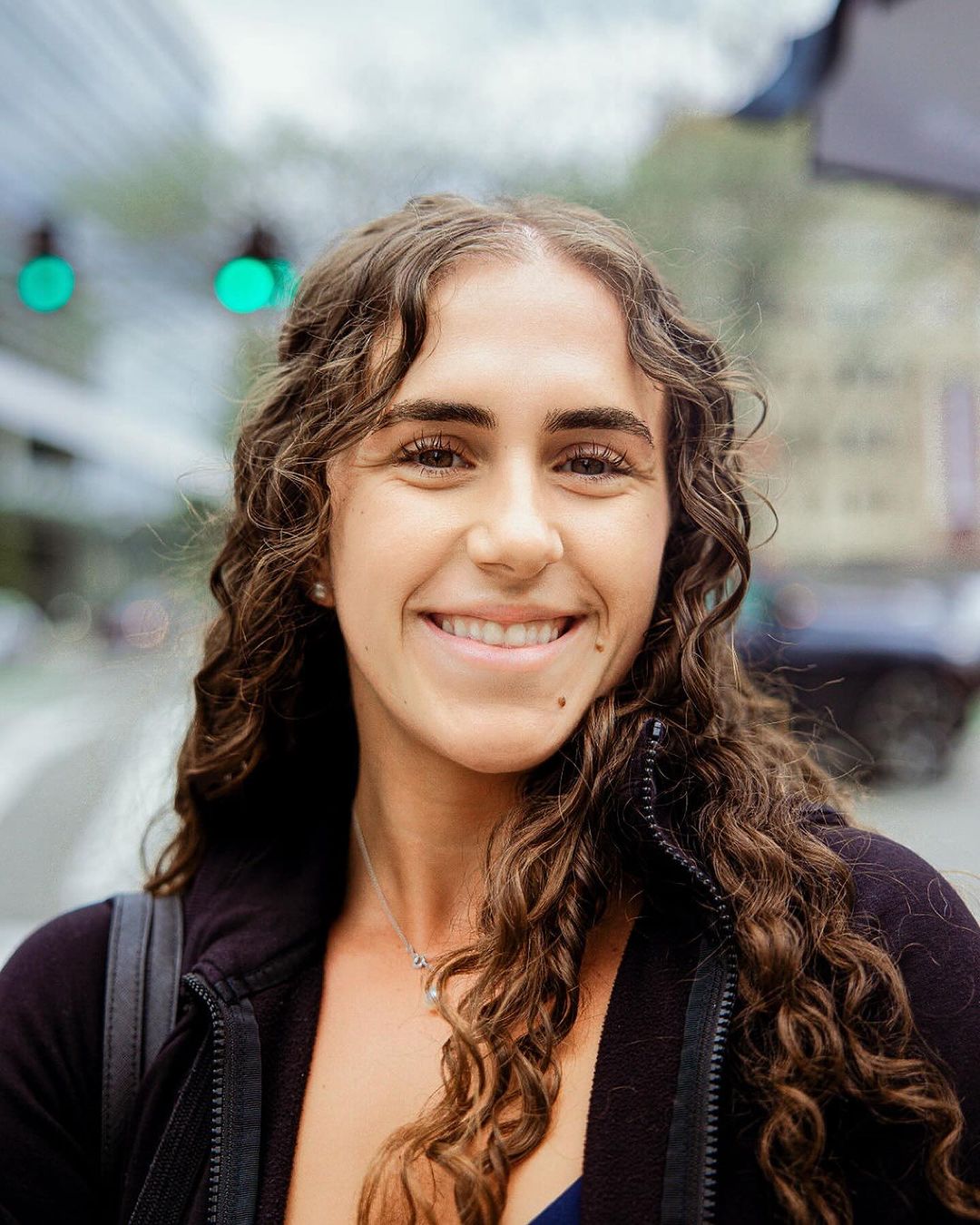 Smiling woman with curly hair stands in a city wearing a black zip-up jacket and a necklace, with traffic lights in the background.