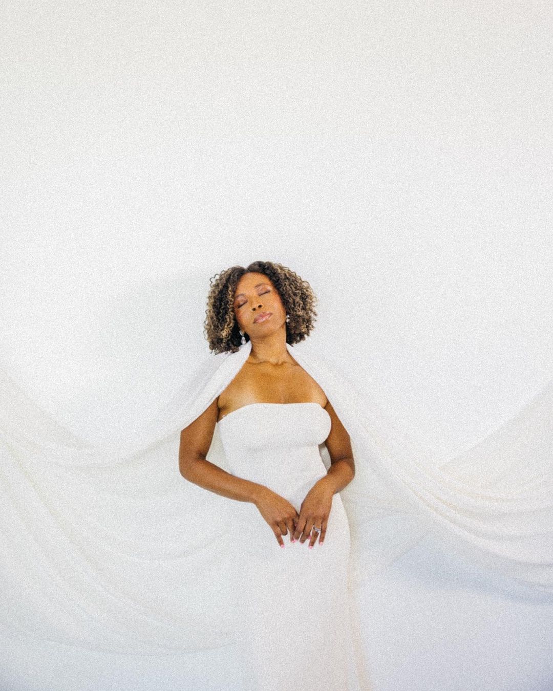 Elegant woman with natural hair in white strapless dress and cape against a minimalist white background in soft lighting. 