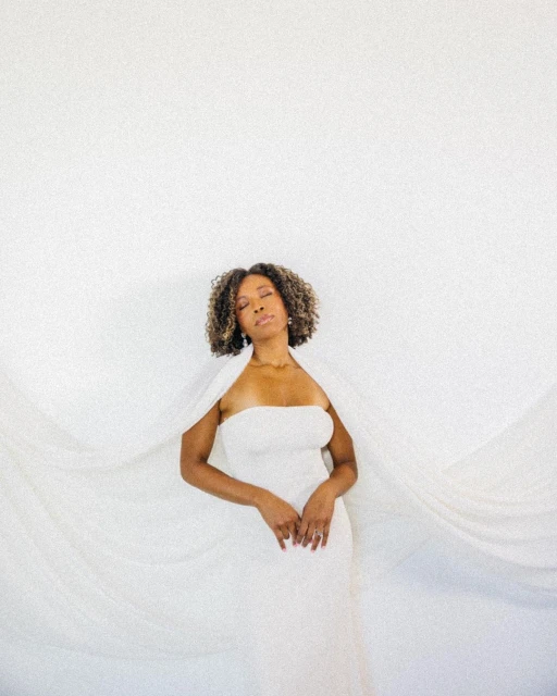 Elegant woman with natural hair in white strapless dress and cape against a minimalist white background in soft lighting. 