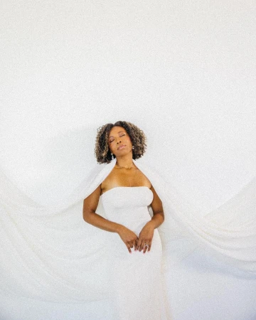 Elegant woman with natural hair in white strapless dress and cape against a minimalist white background in soft lighting. 