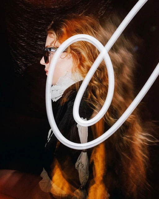 Profile portrait of a red-haired adult woman wearing sunglasses and a ruffled collar, posing in front of a dark background with white neon light.