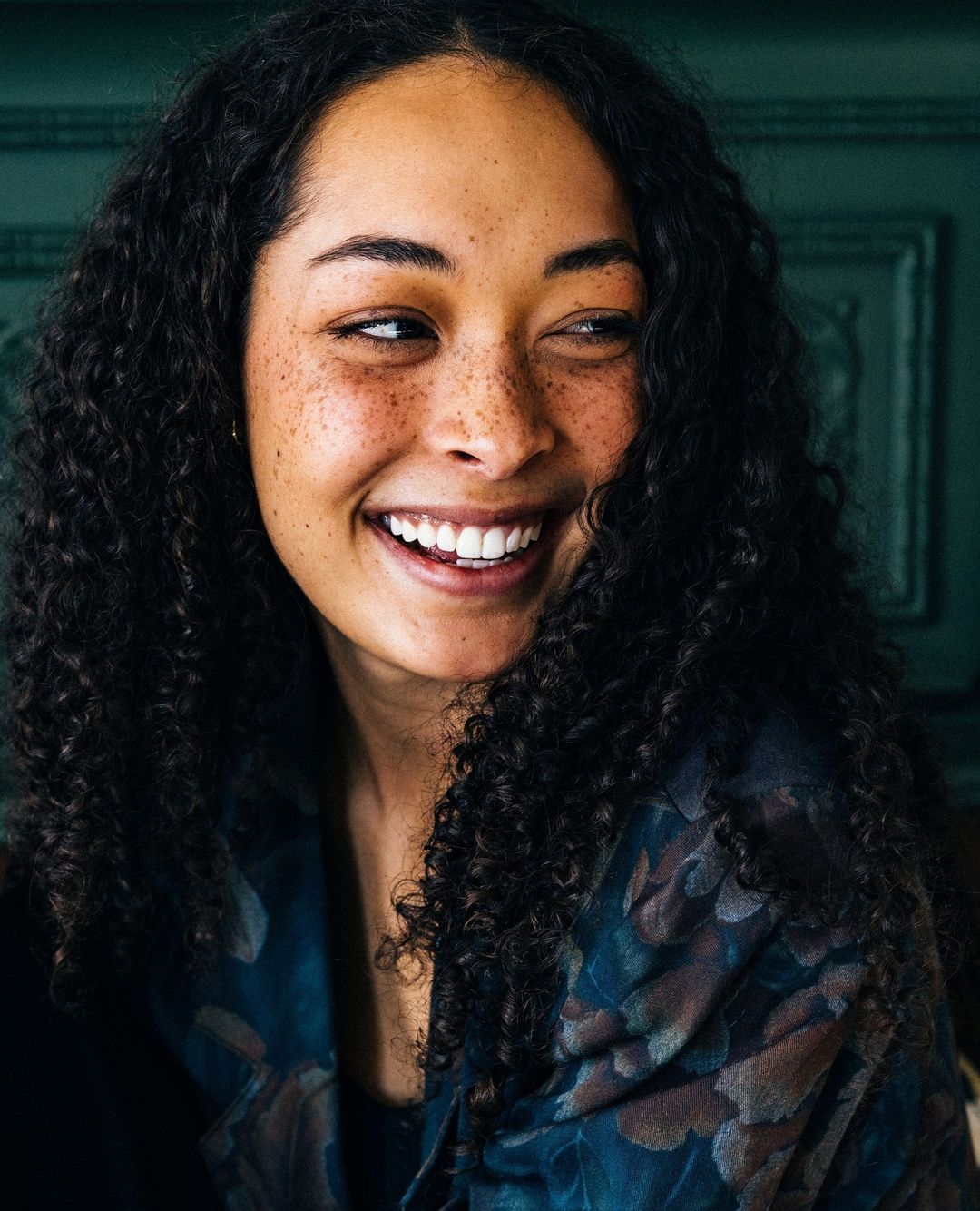 Smiling young woman with curly hair wearing a patterned top, looking to the side against a dark green wall for beauty or fashion.