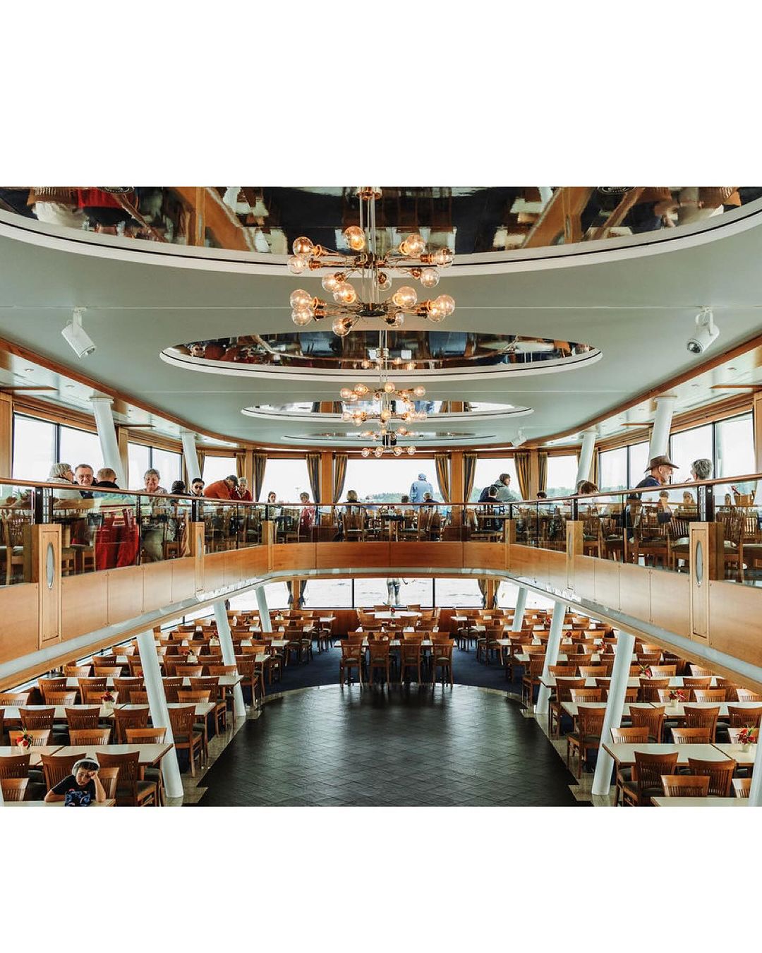 Two-story restaurant interior with rows of tables and chairs, visible patrons on the upper level, and modern chandeliers.