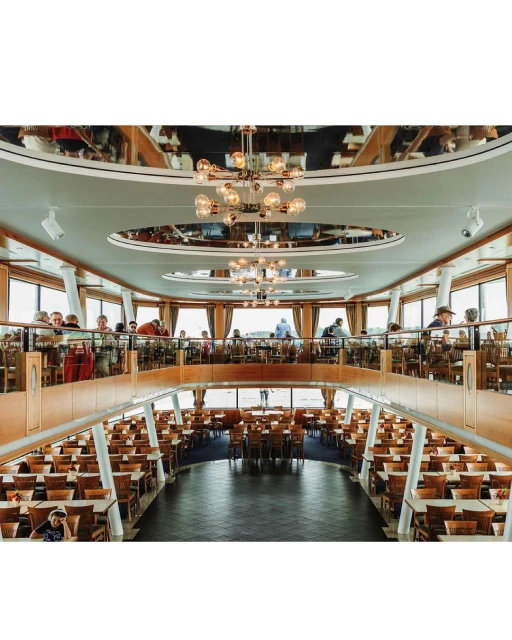 Two-story restaurant interior with rows of tables and chairs, visible patrons on the upper level, and modern chandeliers.