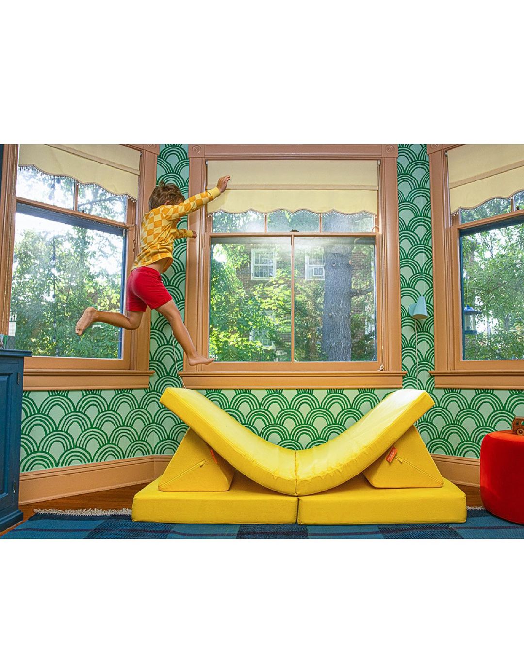 Child jumping through open window above yellow modular furniture, bright interior with green patterned wallpaper, blue floor, and daylight.