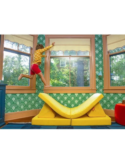Child jumping through open window above yellow modular furniture, bright interior with green patterned wallpaper, blue floor, and daylight.
