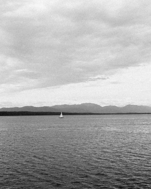 Black and white image of a sailboat on the water, with distant mountains and a cloudy sky in the background, creating a serene scene.