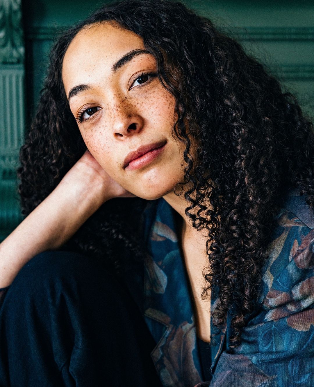 Portrait of a young adult woman with curly hair and freckles, wearing a floral jacket, posing indoors with a serene expression.