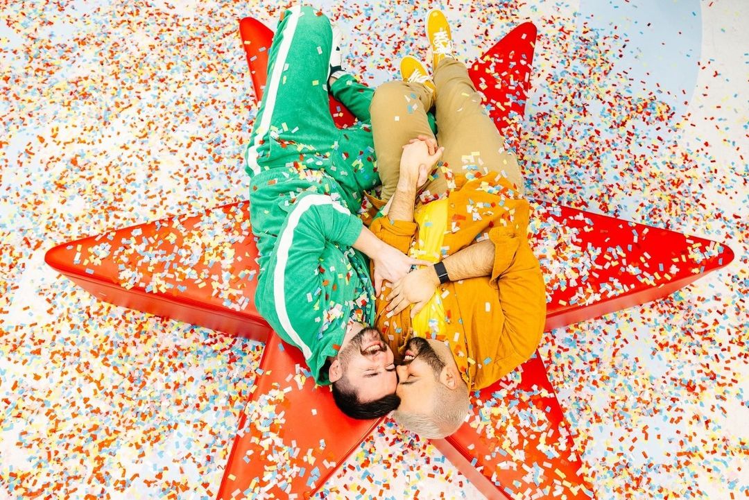 Two adult males in colorful tracksuits lie on a red star-shaped object surrounded by colorful confetti in a brightly lit indoor setting.