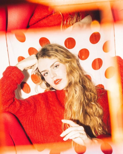 Woman with wavy hair poses indoors wearing a red sweater and red tights against a backdrop with large red polka dots.