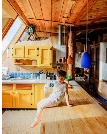 A child sits on a wooden kitchen island in a rustic interior with yellow cabinets and modern hanging lamps, bathed in natural light.