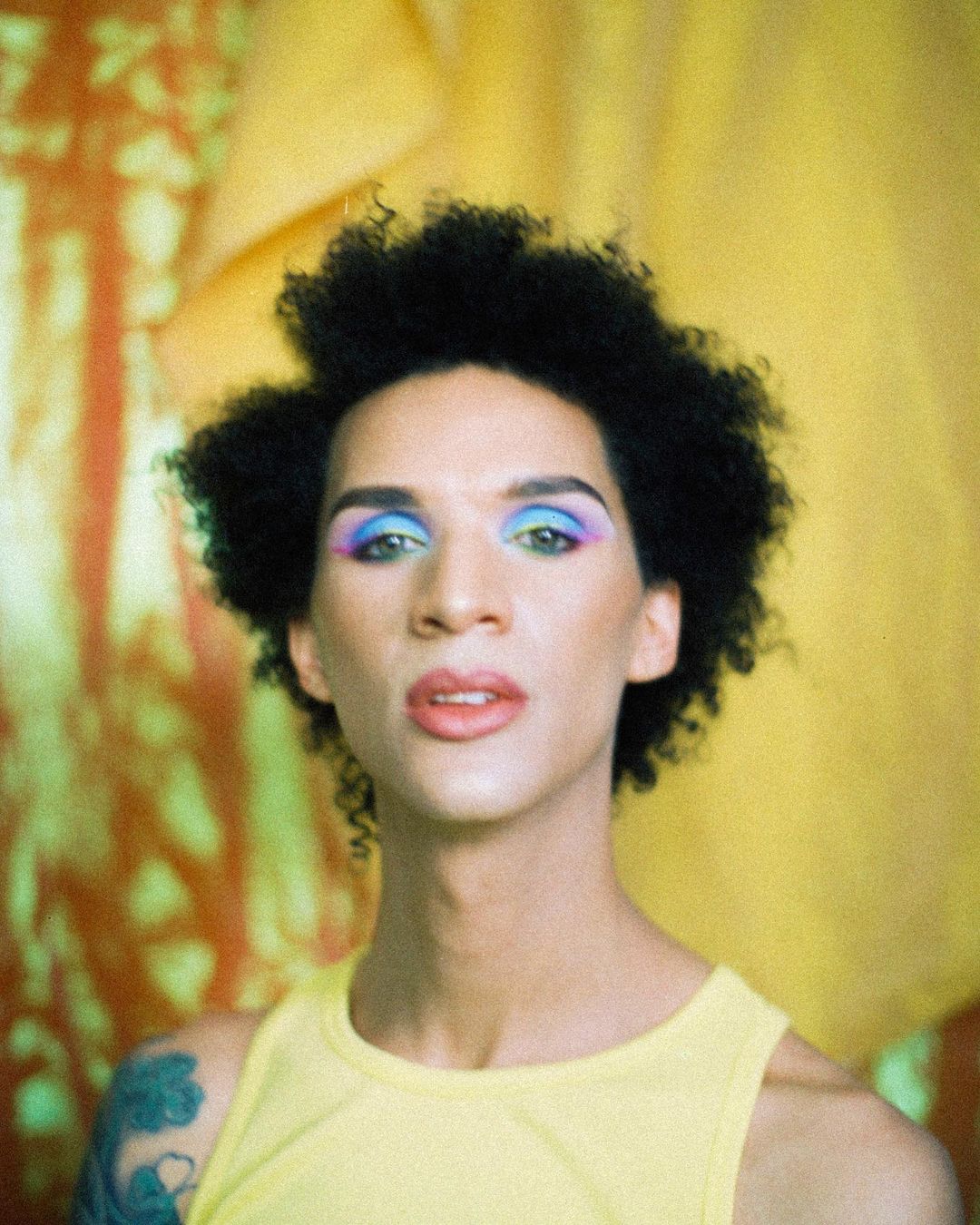Close-up portrait of a person with dark curly hair, wearing makeup and a yellow tank top, posing against a yellow and orange backdrop.
