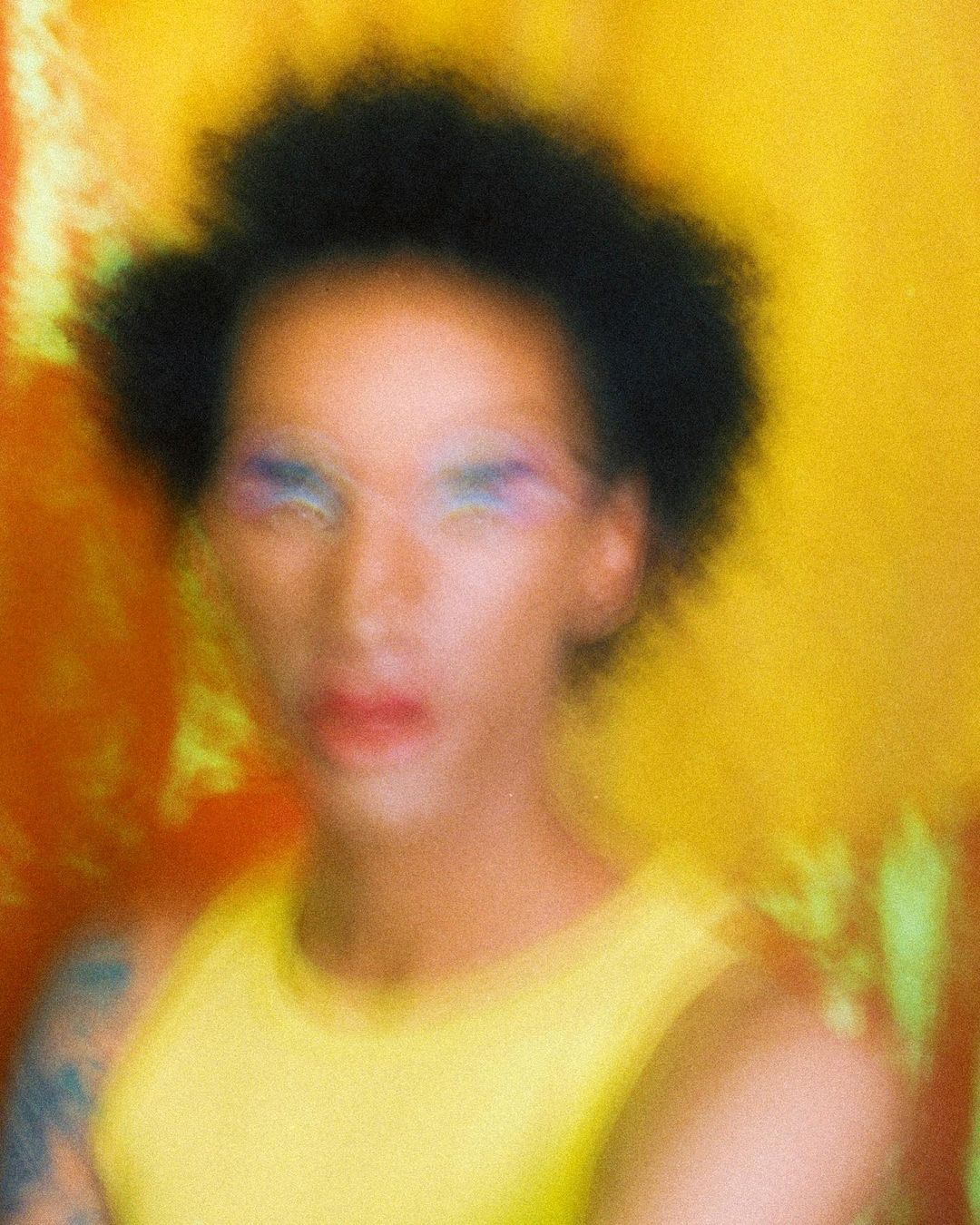 Portrait of an androgynous adult with an afro hairstyle, wearing colorful makeup and a yellow top, against a yellow and orange background.