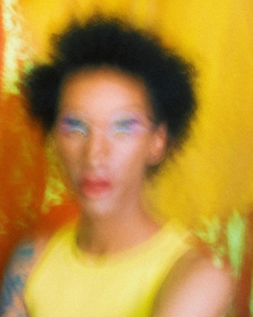 Portrait of an androgynous adult with an afro hairstyle, wearing colorful makeup and a yellow top, against a yellow and orange background.