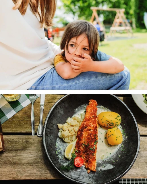 Child rests on a woman's lap outdoors, while a plate of fish with potatoes and lemon sits on a wooden table.