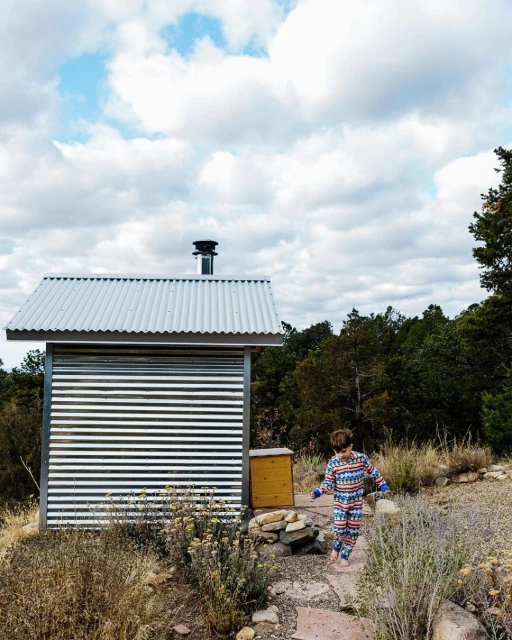 Child in patterned pajamas walks on stone path near small cabin with metal roof, surrounded by vegetation under a cloudy sky.