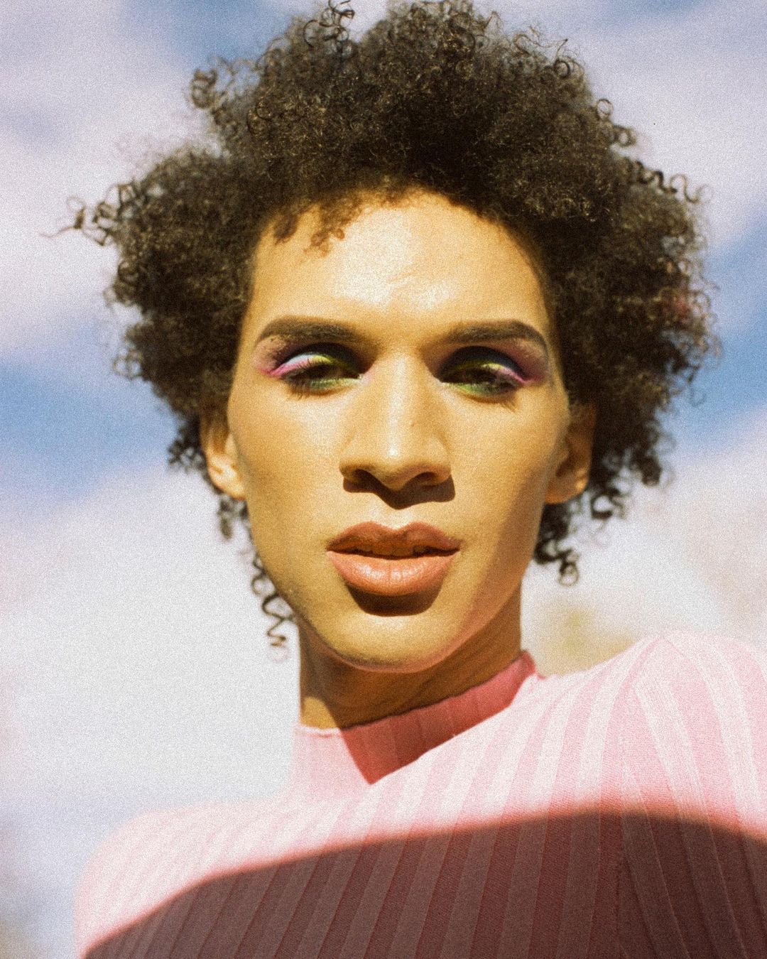 Person with dark curly hair and colourful eye makeup wearing a pink and purple ribbed top, in front of a cloudy blue sky.
